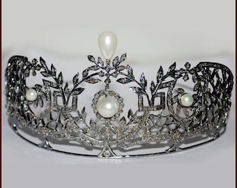 Puede incluir: Una tiara plateada ornamentada con un diseño delicado, con perlas y gemas brillantes. La tiara tiene una gota de perla central y está adornada con motivos de hojas y flores, creando una apariencia elegante y regia.
