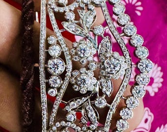 May include: A silver bracelet with a floral design and many small, clear stones. The bracelet is on a pink and white patterned surface.
