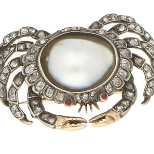 Pode incluir: Um broche de prata e ouro em forma de caranguejo com uma grande pérola no centro e pequenas pedras preciosas vermelhas para os olhos. O caranguejo é adornado com diamantes.