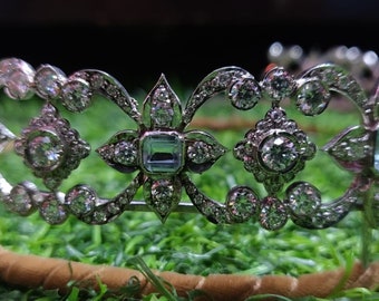 May include: A silver tiara with a delicate design featuring clear and light blue gemstones. The tiara is set on a brown background.