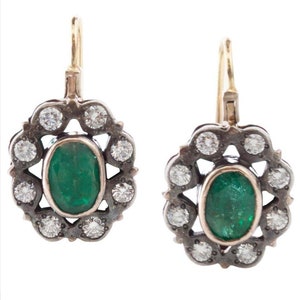 May include: A pair of emerald and diamond earrings with a silver and gold setting. The earrings have a floral design with a large oval emerald in the centre and smaller diamonds surrounding it.