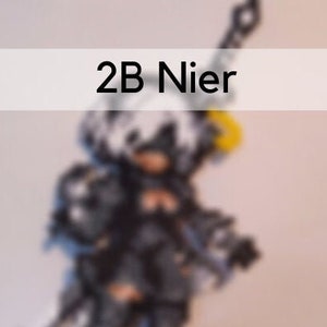 May include: A figurine of the character 2B from Nier, in a black and silver outfit, holding a sword and a weapon. The figure has a white hair and a yellow accessory. The text "2B Nier" is displayed above the figure.
