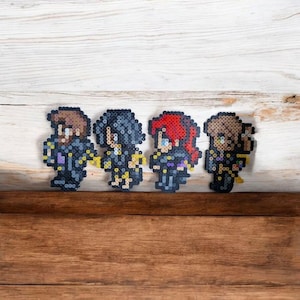Peut inclure: Quatre figurines en pixel art de personnages aux cheveux bruns, rouges et noirs, vêtus de tenues noires et jaunes. Les figurines sont disposées sur une surface en bois avec un fond blanc et marron. Les figurines sont faites de petites perles colorées.