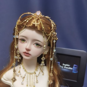 May include: A doll with long brown hair wearing a gold headpiece featuring a central pink stone, dangling chains, and small crystals. The doll also wears a matching gold necklace with white beads and hanging chains, and small gold stud earrings.