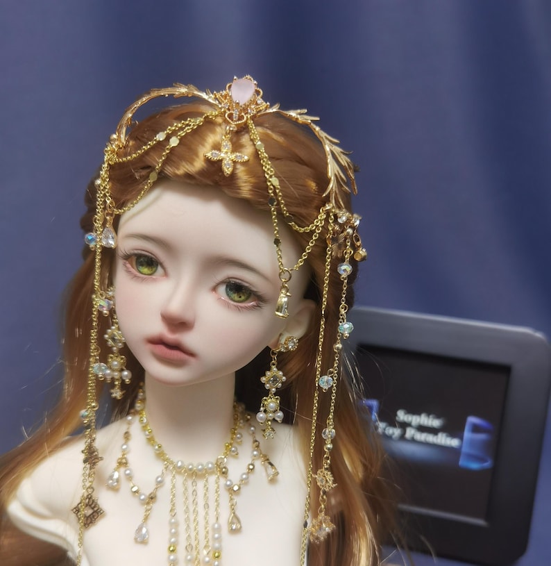 May include: A doll with long brown hair wearing a gold headpiece featuring a central pink stone, dangling chains, and small crystals. The doll also wears a matching gold necklace with white beads and hanging chains, and small gold stud earrings.
