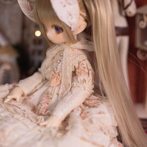 May include: A porcelain doll with blonde hair styled in pigtails, wearing a white dress with floral print and a white lace bonnet.
