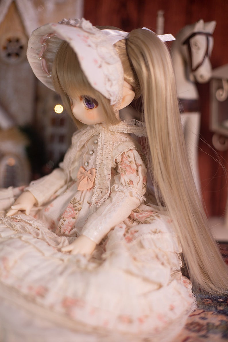 May include: A porcelain doll with blonde hair styled in pigtails, wearing a white dress with floral print and a white lace bonnet.