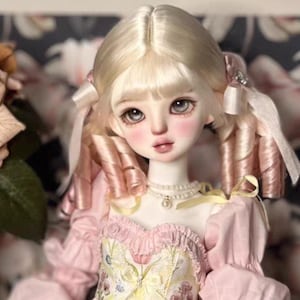 May include: A ball-jointed doll with pale skin, large grey eyes, and long blonde hair styled in pigtails with pink ribbons. The doll wears a pearl necklace and a pink dress featuring a floral pattern on the bodice and puffed sleeves.