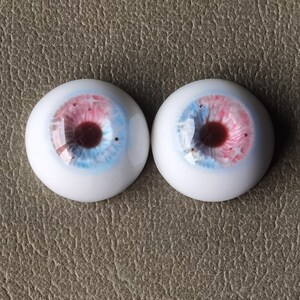 May include: A pair of blue, pink, and brown doll eyes with a white iris. The eyes are made of glass and have a realistic appearance.