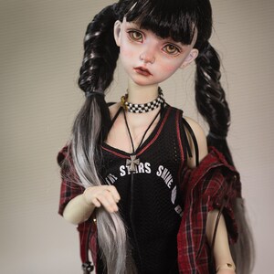 Puede incluir: Una mu&ntilde;eca con el pelo largo trenzado en negro y blanco, lleva un top negro sin mangas con el texto "All Stars Shine Bright" y una camisa de cuadros rojos y negros. La mu&ntilde;eca lleva un collar con una cruz plateada y una gargantilla negra y blanca a cuadros.