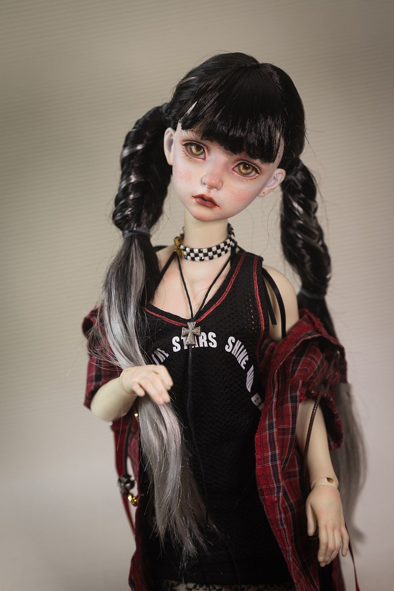 Puede incluir: Una mu&ntilde;eca con el pelo largo trenzado en negro y blanco, lleva un top negro sin mangas con el texto "All Stars Shine Bright" y una camisa de cuadros rojos y negros. La mu&ntilde;eca lleva un collar con una cruz plateada y una gargantilla negra y blanca a cuadros.