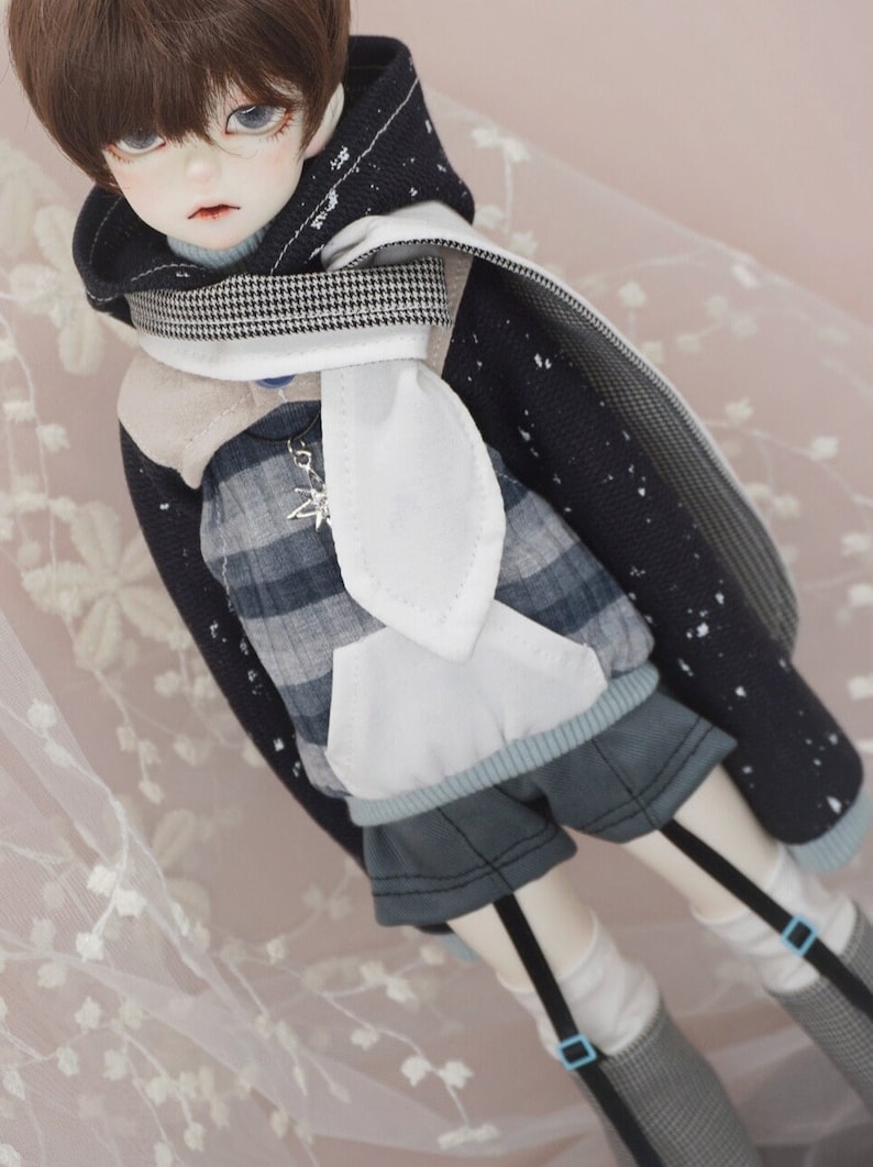 May include: A doll wearing a blue and white plaid shirt, a white hooded sweater with a black and white checkered scarf, and blue shorts with black suspenders.