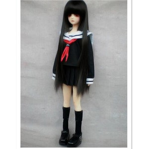 May include: A black doll with long black hair wearing a black and white sailor uniform with a red bow tie. The doll is wearing black socks and black shoes.