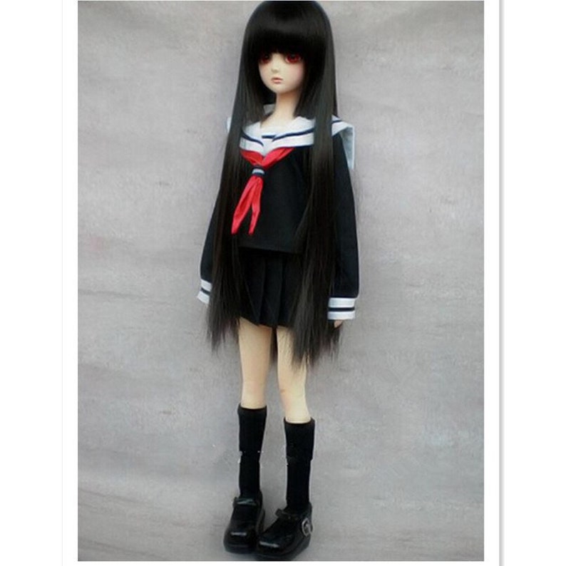 May include: A black doll with long black hair wearing a black and white sailor uniform with a red bow tie. The doll is wearing black socks and black shoes.