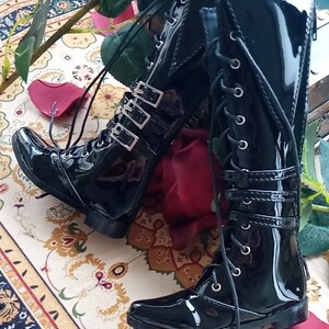 May include: A pair of black patent leather knee-high boots with buckles and laces. The boots have a pointed toe and a low heel.