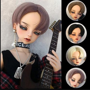 May include: A doll with a short, straight, light brown wig with a side part. The doll is wearing a black shirt, a silver chain necklace with a dog tag, and a black choker with the word "NICE" on it. The doll is holding a guitar.