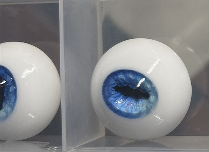 May include: A pair of blue and white doll eyes with a white sclera and a blue iris. The eyes are displayed in a clear plastic container.