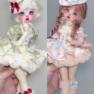 May include: Two ball-jointed dolls are shown, each with a unique style. One doll wears a green dress and a hat with a cherry on top. The other doll is dressed in pink and has a similar hat. Both dolls have detailed facial features and are wearing shoes.