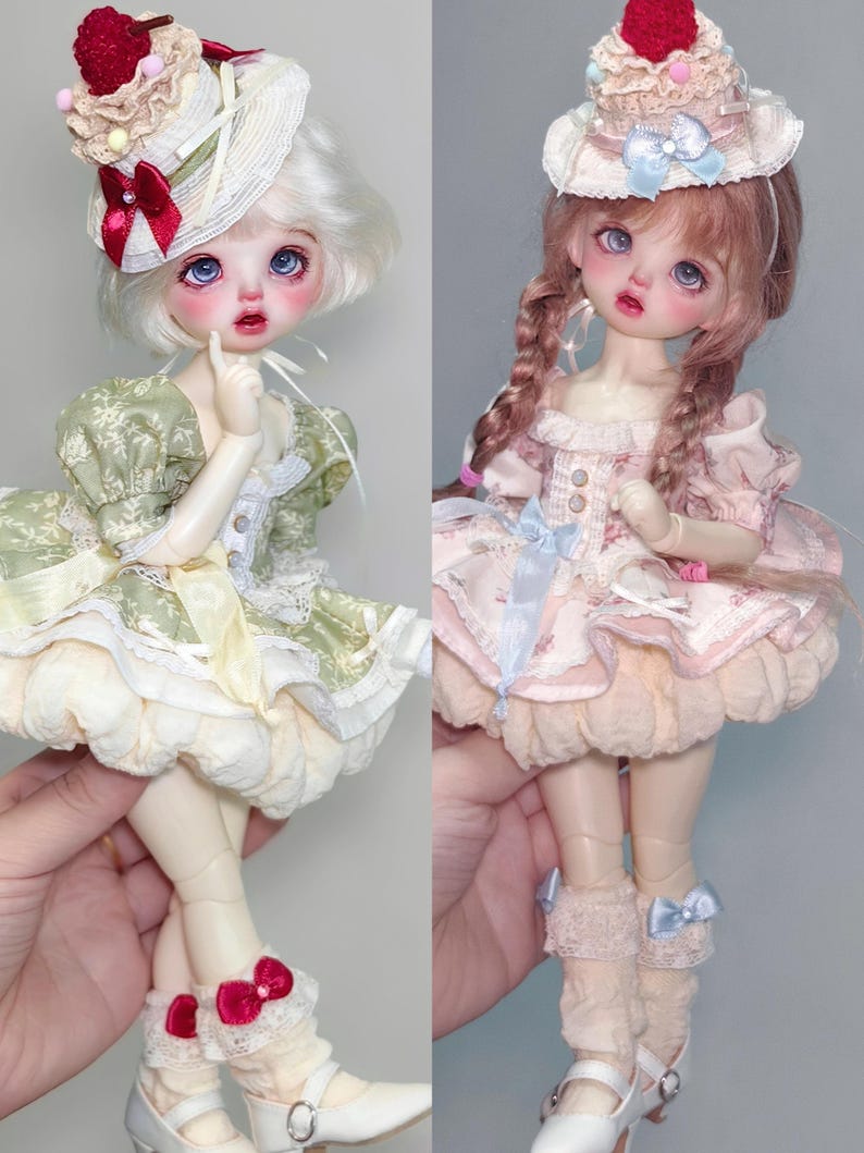 May include: Two ball-jointed dolls are shown, each with a unique style. One doll wears a green dress and a hat with a cherry on top. The other doll is dressed in pink and has a similar hat. Both dolls have detailed facial features and are wearing shoes.