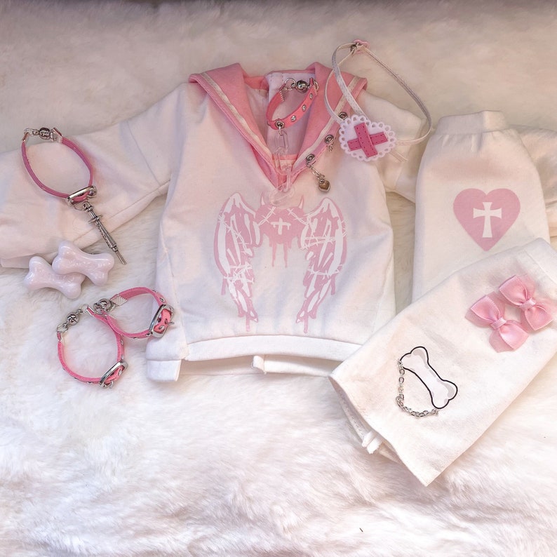 K&ouml;nnte beinhalten: Ein wei&szlig;es und rosafarbenes Outfit mit einem Matrosenkragen, einem rosafarbenen Herzen mit einem Kreuz und einem rosafarbenen Engelsfl&uuml;gel-Design. Das Outfit beinhaltet ein lang&auml;rmliges Shirt, eine Hose und ein Stirnband. Das Outfit beinhaltet auch ein rosafarbenes Halsband, einen rosafarbenen knochenf&ouml;rmigen Anh&auml;nger und eine rosafarbene Kette.