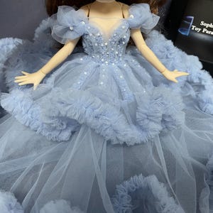 May include: A light blue tulle dress with a ruffled skirt and a beaded bodice. The dress has a sweetheart neckline and off-the-shoulder straps. The dress is decorated with small white beads.