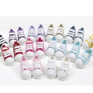 Puede incluir: Una colección de 20 zapatillas de lona en miniatura de varios colores, incluyendo negro, blanco, rojo, rosa, morado, azul, amarillo y beige. Las zapatillas tienen cordones blancos y están dispuestas en filas sobre una superficie blanca.