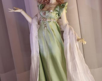 May include: A ball-jointed doll with wavy light brown hair wearing a pale green satin empire-waist gown with puff sleeves. The doll features a white floral headpiece, layered pearl necklaces, and a long, sheer white striped shawl draped over the arms.