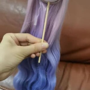 Puede incluir: Una peluca de muñeca con un degradado de colores, que va del morado claro al azul. La peluca está sujeta a una cabeza blanca y se sujeta con un palito de madera. El pelo es largo y ondulado, con una textura suave y fluida.