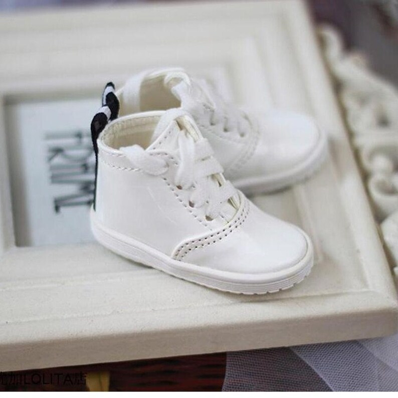 May include: A pair of white leather sneakers with black ribbon laces. The sneakers have a white rubber sole and are tied with a black bow.