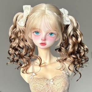 May include: A doll with a pale complexion, rosy cheeks, and bright blue eyes. The doll has blonde hair styled in pigtails with white bows. The hair transitions to a darker brown at the ends. The doll is wearing a beige lace top.