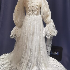 May include: A white lace dress with long sleeves and a high neckline. The dress has a fitted bodice and a flowing skirt. The dress is decorated with lace and buttons. The dress is on a mannequin.