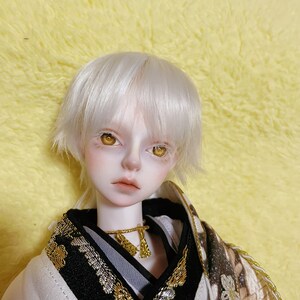 Puede incluir: Una mu&ntilde;eca de pelo blanco con ojos dorados que lleva un kimono estampado en blanco y negro con detalles dorados.