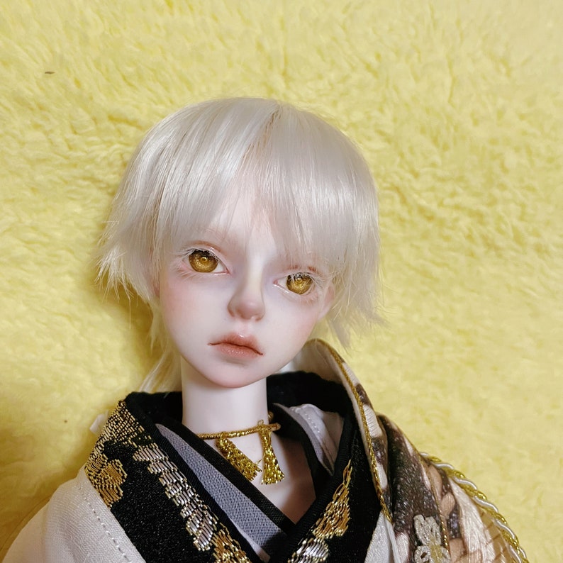 Puede incluir: Una mu&ntilde;eca de pelo blanco con ojos dorados que lleva un kimono estampado en blanco y negro con detalles dorados.
