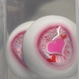 May include: Two white doll eyes with pink and silver glitter accents. Each eye has a pink heart design with smaller red hearts.