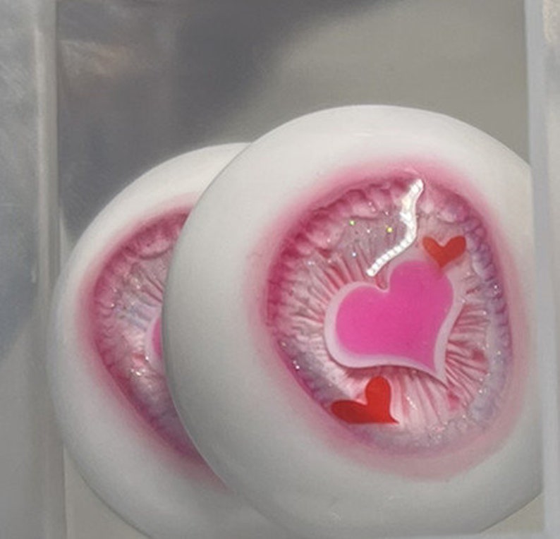 May include: Two white doll eyes with pink and silver glitter accents. Each eye has a pink heart design with smaller red hearts.