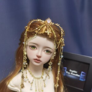 May include: A doll with long auburn hair wearing a gold headpiece featuring a central pink stone and dangling chains. The doll has green eyes and is adorned with a matching gold necklace featuring small white pearls and clear crystals.