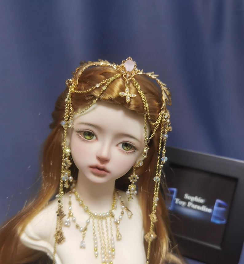 May include: A doll with long auburn hair wearing a gold headpiece featuring a central pink stone and dangling chains. The doll has green eyes and is adorned with a matching gold necklace featuring small white pearls and clear crystals.