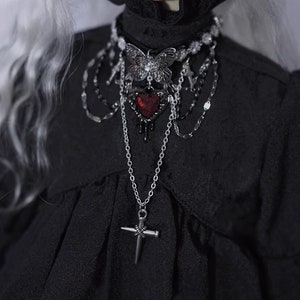 K&ouml;nnte beinhalten: Eine silberne und schwarze Choker-Kette mit einem Schmetterlingsanh&auml;nger, einem roten Herzanh&auml;nger und einem Kreuzanh&auml;nger. Die Kette wird von einer Puppe mit wei&szlig;en Haaren und einem schwarzen Kleid getragen.