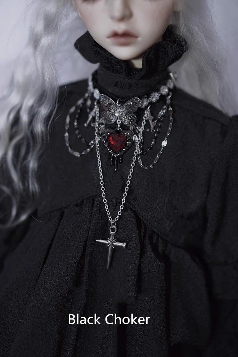 K&ouml;nnte beinhalten: Eine silberne und schwarze Choker-Kette mit einem Schmetterlingsanh&auml;nger, einem roten Herzanh&auml;nger und einem Kreuzanh&auml;nger. Die Kette wird von einer Puppe mit wei&szlig;en Haaren und einem schwarzen Kleid getragen.