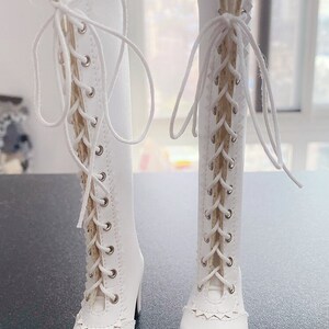 Pode incluir: Um par de botas brancas at&eacute; ao joelho com detalhes de atacadores. As botas apresentam um salto alto e um design de borda ondulada &agrave; volta do tornozelo. A parte superior da bota &eacute; adornada com renda delicada.