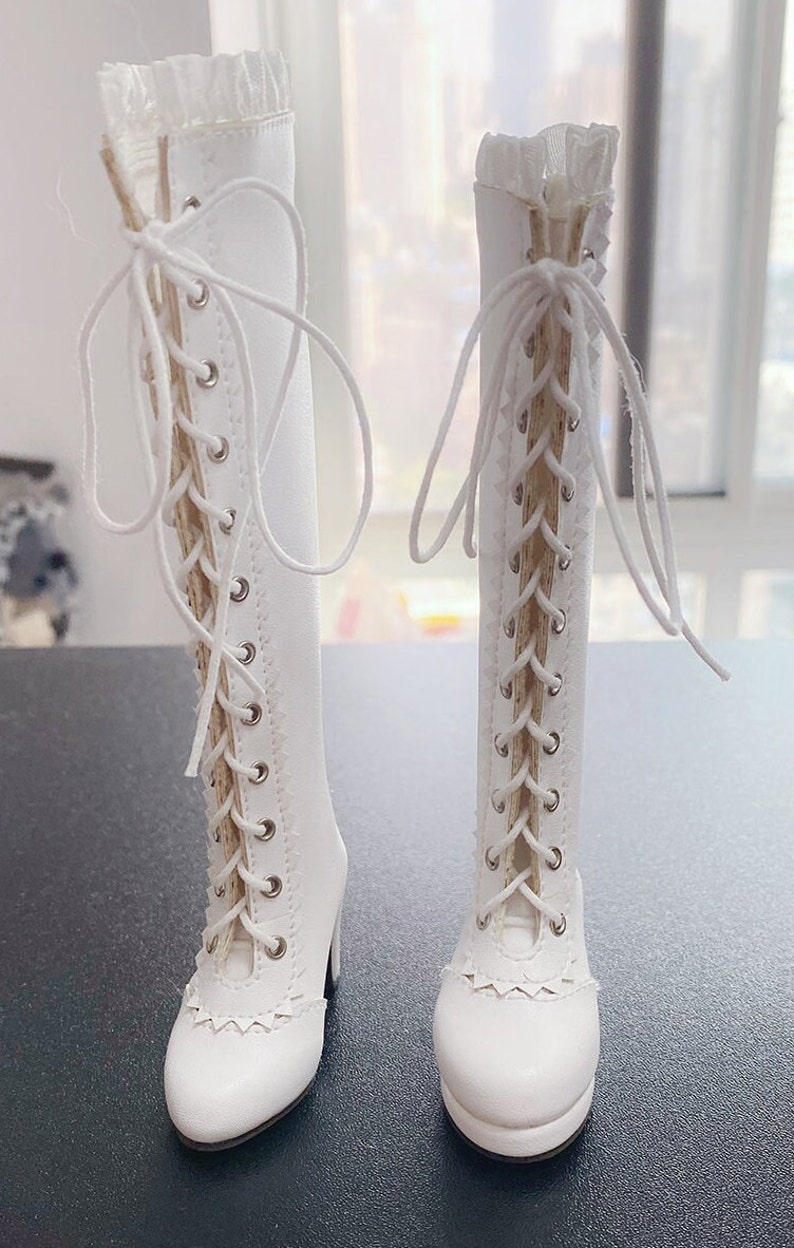 Pode incluir: Um par de botas brancas at&eacute; ao joelho com detalhes de atacadores. As botas apresentam um salto alto e um design de borda ondulada &agrave; volta do tornozelo. A parte superior da bota &eacute; adornada com renda delicada.