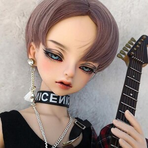 May include: A doll with short, light brown hair, wearing a black choker with the word "NICE" on it, a silver chain necklace, and a black shirt. The doll is holding a guitar and has blue eyes.