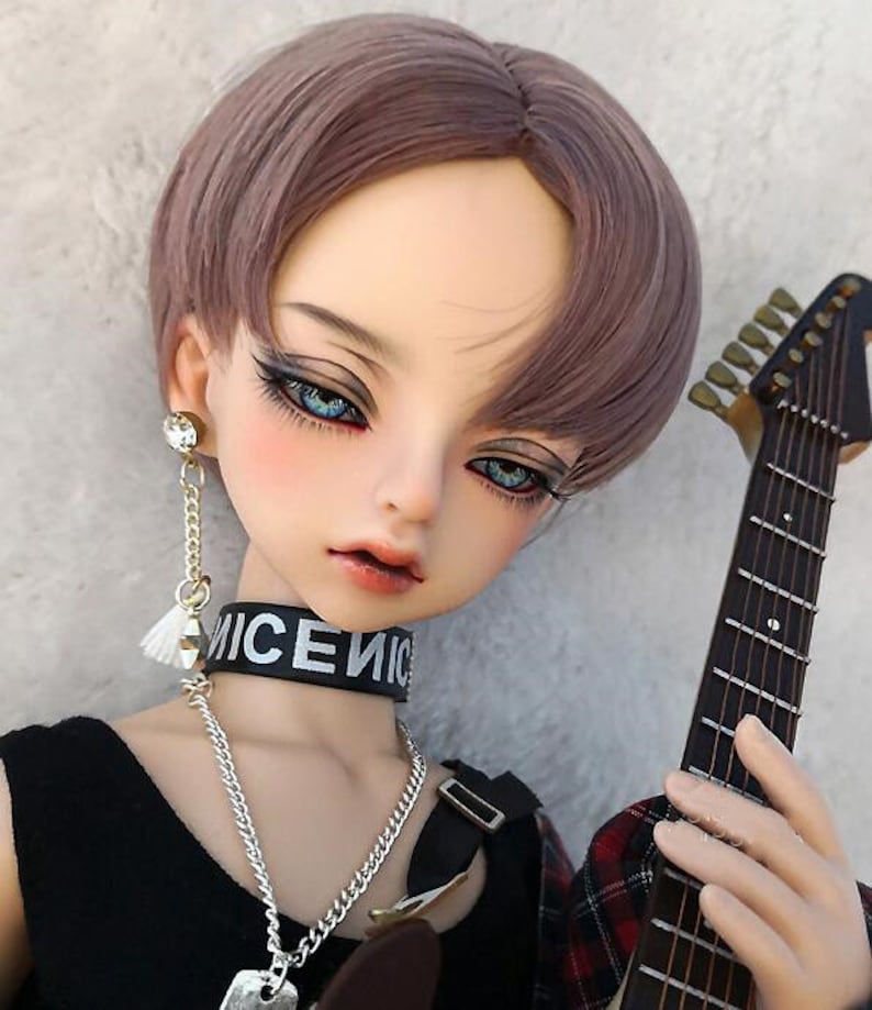 May include: A doll with short, light brown hair, wearing a black choker with the word "NICE" on it, a silver chain necklace, and a black shirt. The doll is holding a guitar and has blue eyes.
