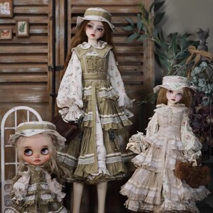 May include: Three dolls wearing vintage-style dresses and hats. The dolls are standing in front of a wooden background. The doll on the left is wearing a green and white dress with a green hat. The doll in the middle is wearing a green and white dress with a brown hat. The doll on the right is wearing a beige and white dress with a white hat.