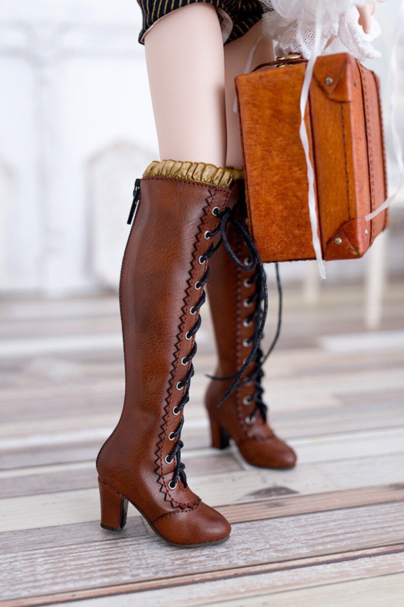 Pode incluir: Botas castanhas at&eacute; ao joelho com atacadores pretos e salto pequeno. As botas t&ecirc;m um padr&atilde;o de costura decorativo nas laterais e uma borda dourada. Uma pequena mala castanha &eacute; segurada perto. Altura das botas: aprox. 30 cm.