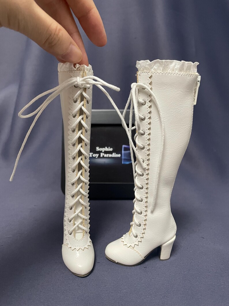 Pode incluir: Um par de botas brancas para bonecas com detalhes de cadar&ccedil;os e saltos pequenos. As botas t&ecirc;m uma borda ondulada na parte superior e s&atilde;o amarradas com cadar&ccedil;os brancos. As botas s&atilde;o projetadas para uma boneca.