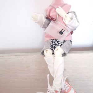 May include: A doll wearing a pink and grey outfit with a white shirt, a pink sweater, and a black and white patterned skirt. The doll is holding a pink purse with a bunny charm. The doll is wearing white socks and pink and white sneakers.