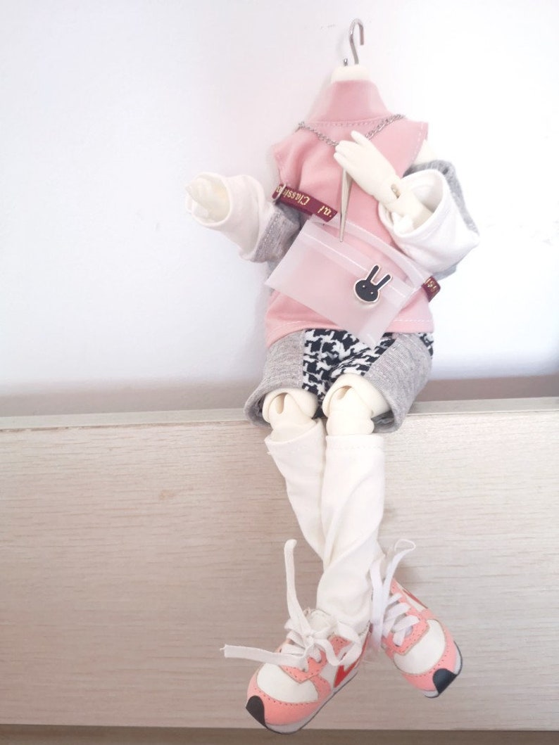May include: A doll wearing a pink and grey outfit with a white shirt, a pink sweater, and a black and white patterned skirt. The doll is holding a pink purse with a bunny charm. The doll is wearing white socks and pink and white sneakers.