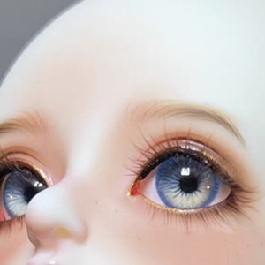 May include: Close-up of a doll's face with blue eyes and long eyelashes. The doll's skin is pale and smooth.