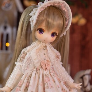 May include: A doll with long blonde hair styled in pigtails, wearing a white dress with pink floral accents and a white lace bonnet with pink trim.